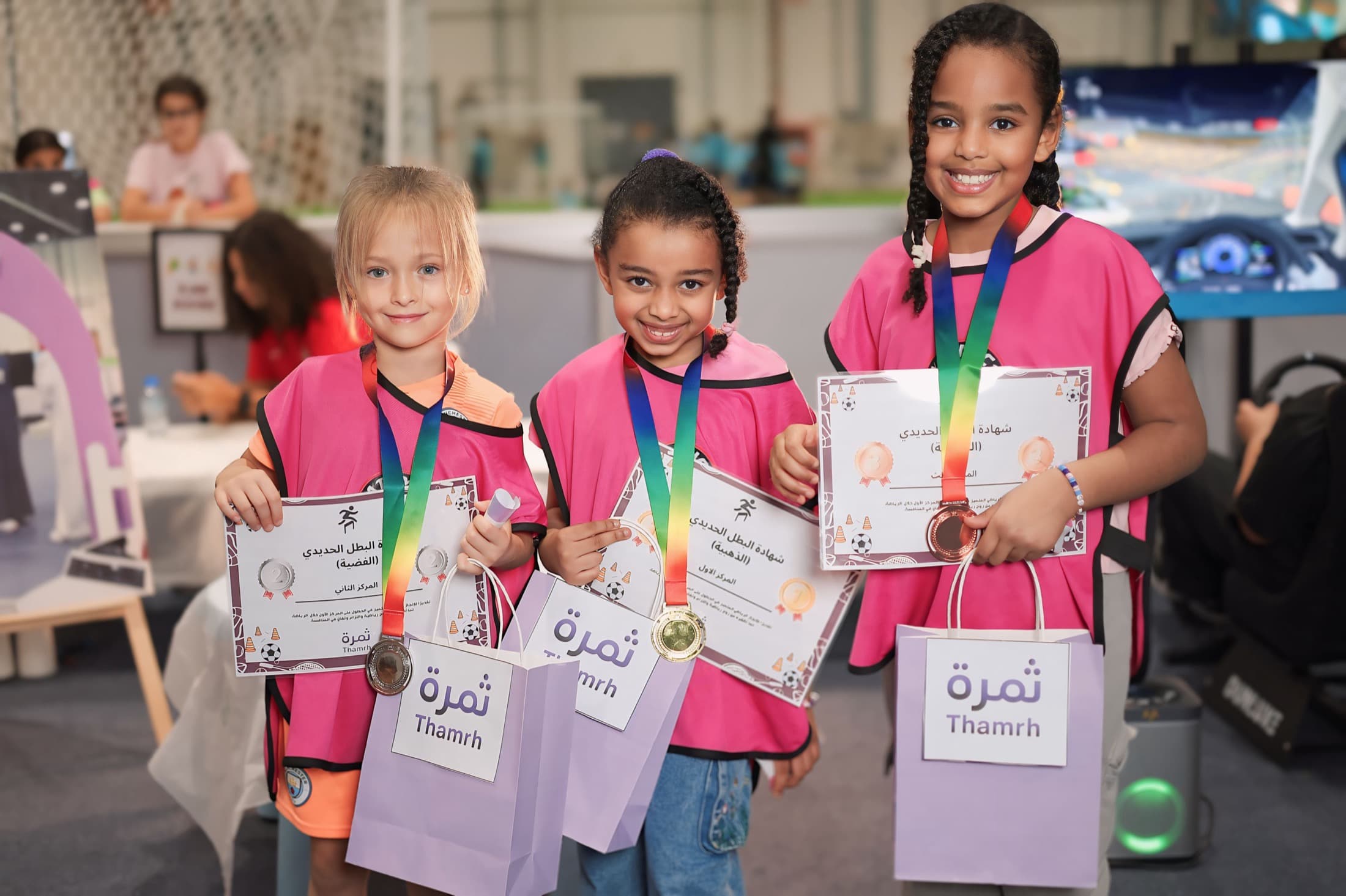 Three girls proudly holding medals, certificates, and Thamrh gift bags